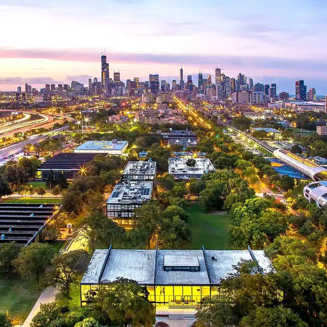 Aerial view of Illinois Tech's campus at dusk, with S.R. Crown Hall lit up in front and the Chicago skyline behind it.