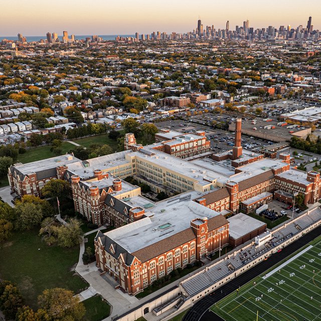 Aerial view of Lane Tech's red-brick campus and football field, with the Chicago skyline behind it.