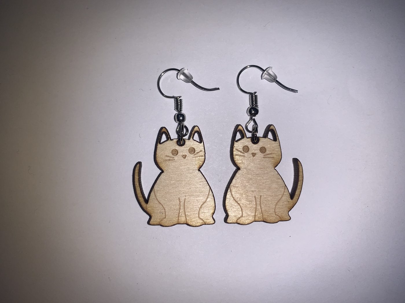 A pair of laser-cut wooden sitting-cat earrings with engraved faces, on a white background