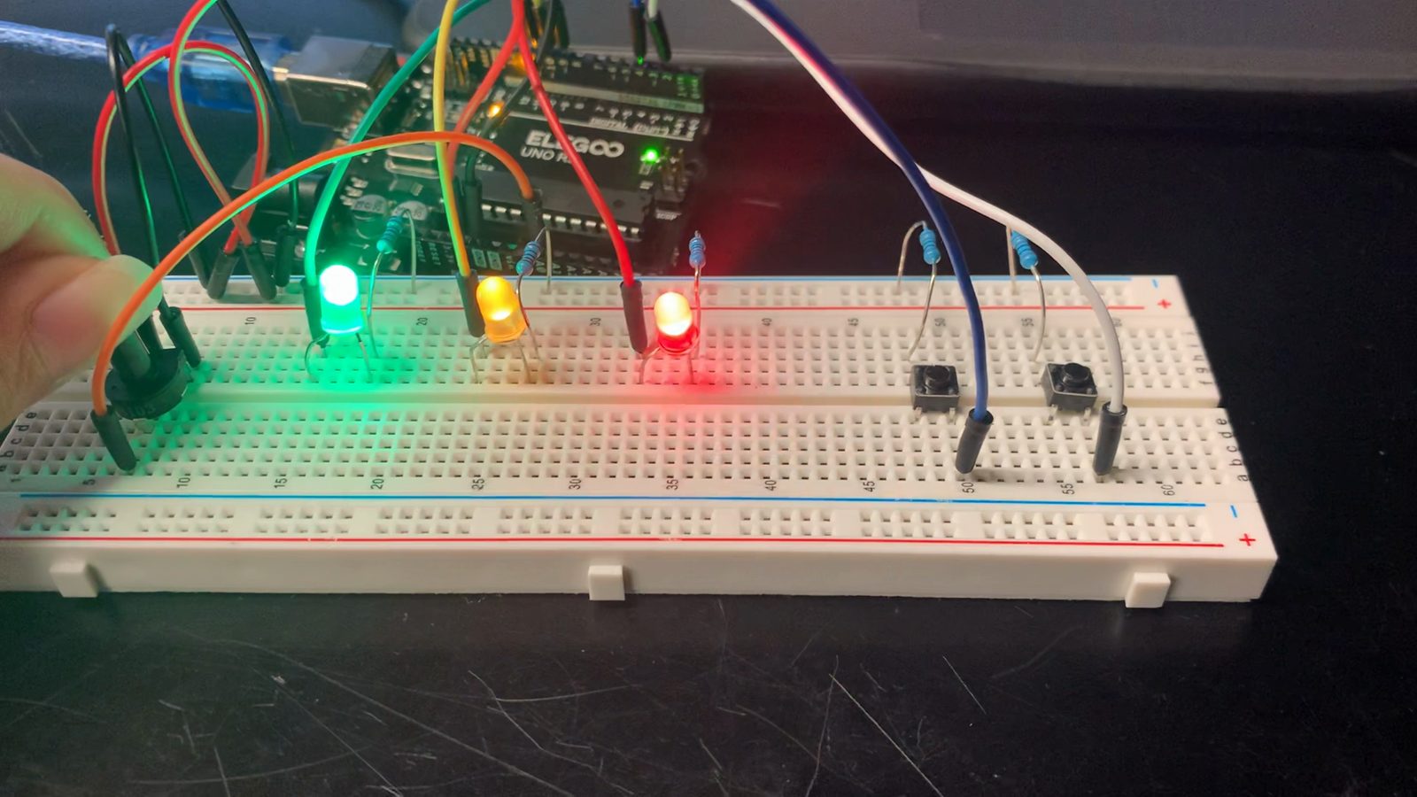 A breadboard with green, yellow and red LEDs all lit, a hand at the black potentiometer knob, two push buttons on the right and jumpers running to an Arduino.