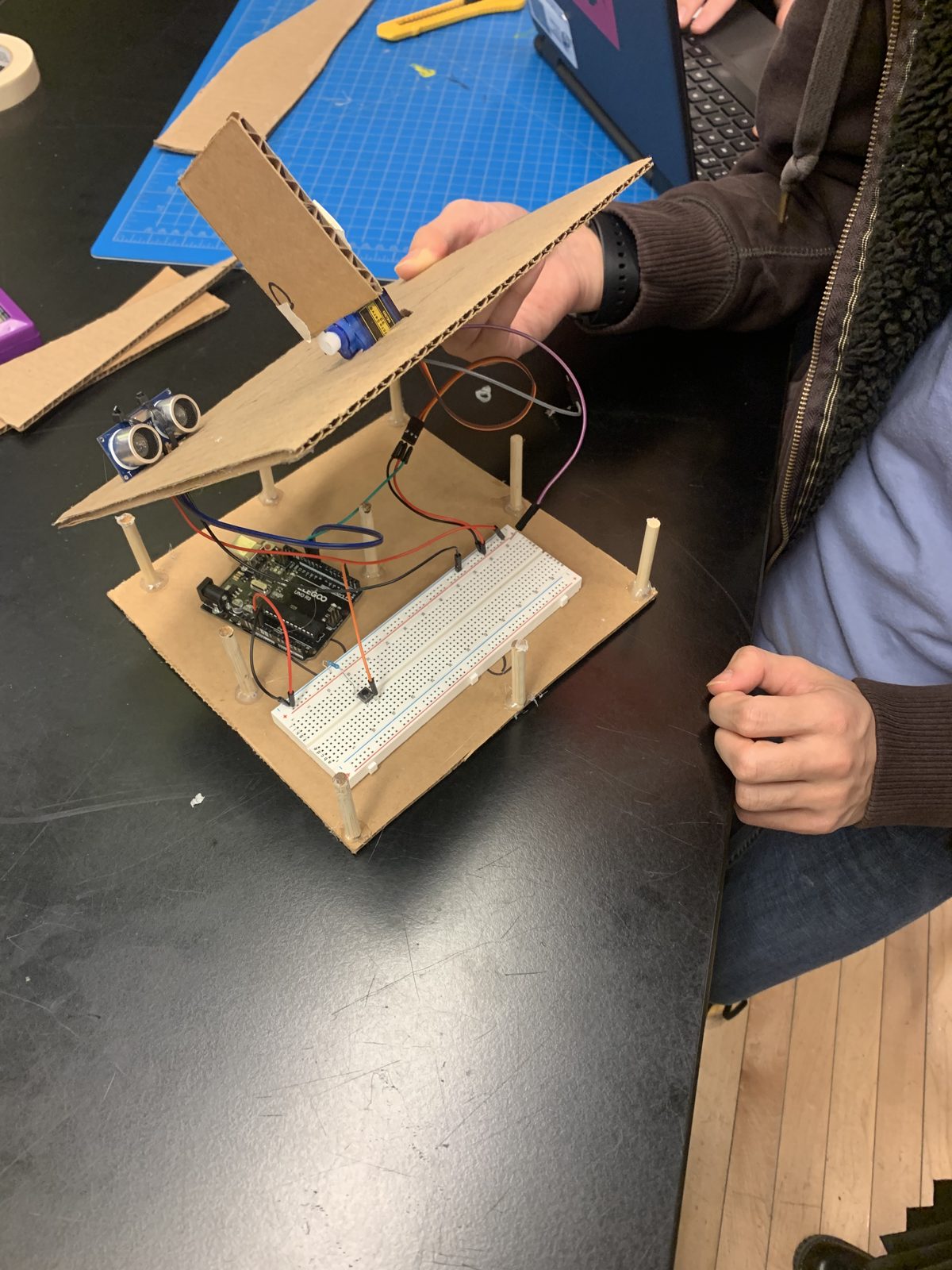 A two-level model elevator made of cardboard platforms on wooden dowels, with an Arduino, breadboard and ultrasonic sensor mounted on it.